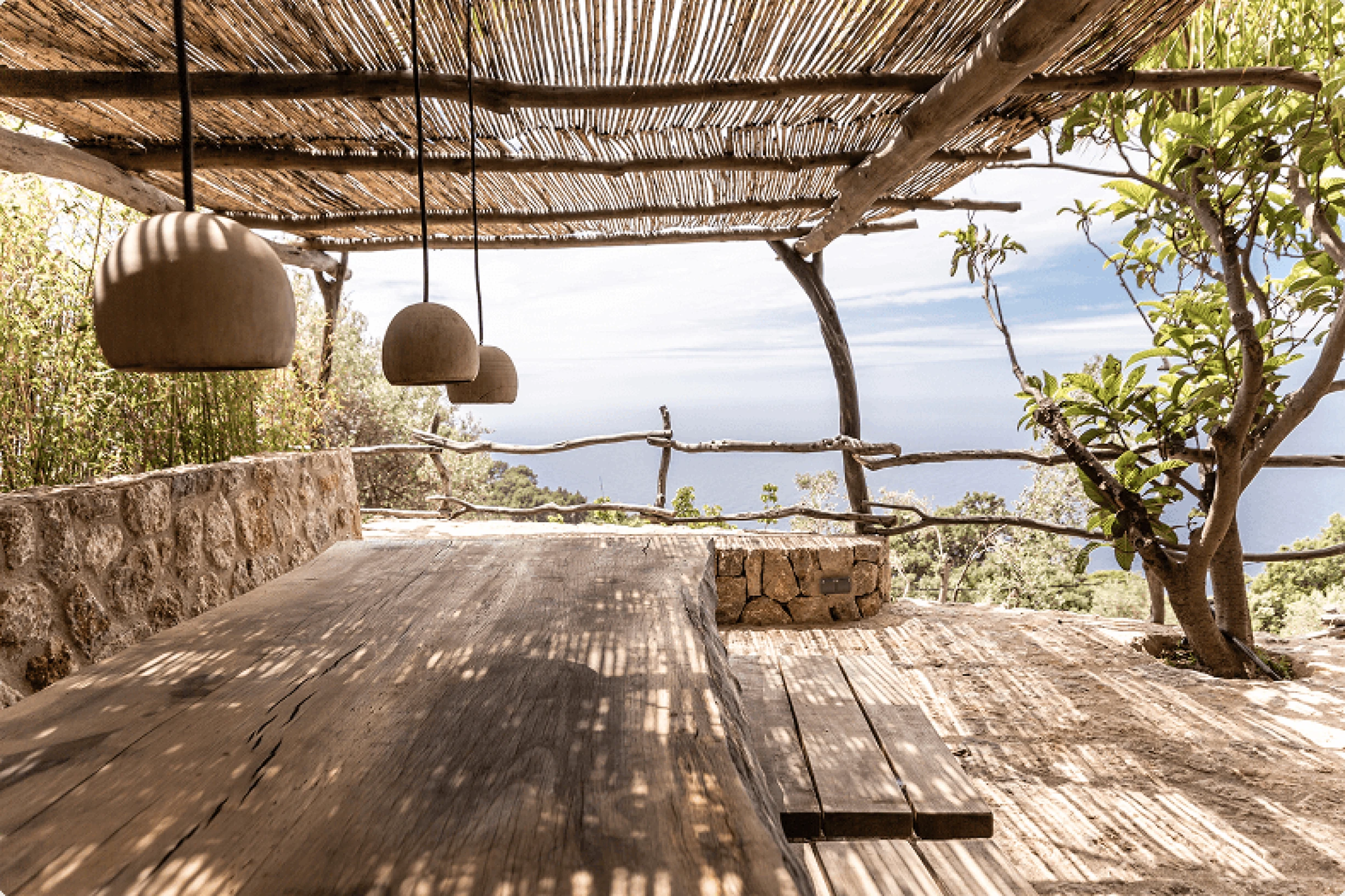 Rustic outdoor dining terrace featuring a shaded pergola, traditional dry stone walls, and panoramic Mediterranean sea views
